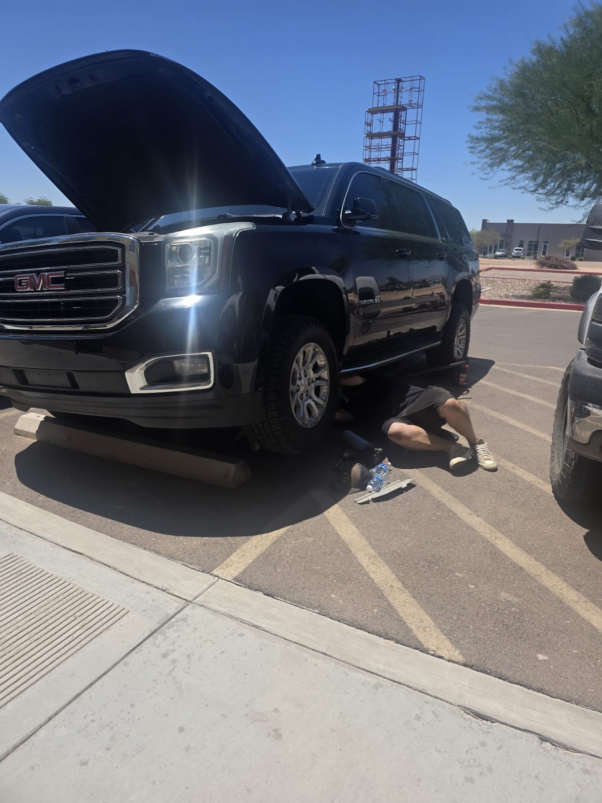 Working underneath a black GMC Yukon XL with the hood up, in a sunny parking lot
